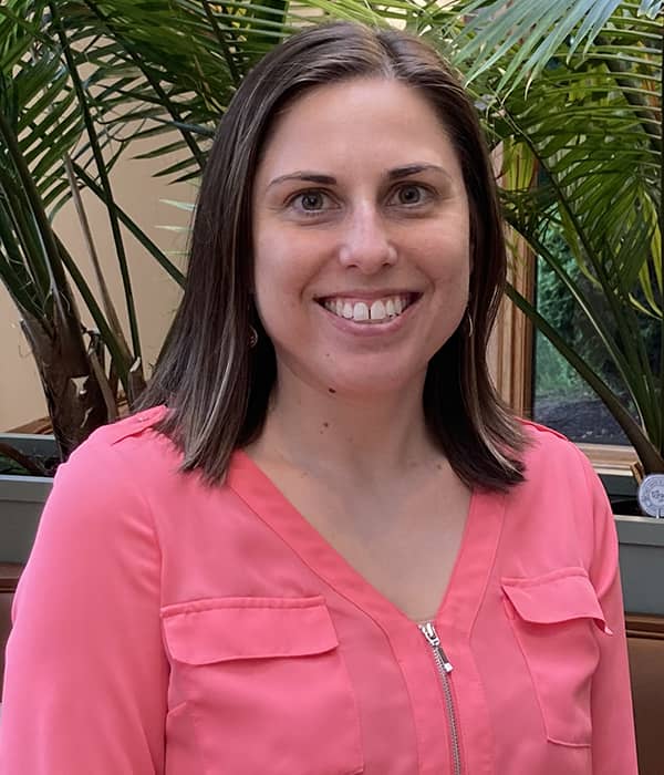 Dr. Jacquelyn Boyd, a smiling woman with shoulder-length brown hair, is wearing a bright pink blouse. She is seated in front of a vibrant arrangement of green plants, giving the setting a fresh and friendly atmosphere.