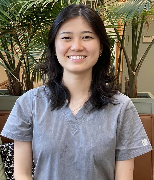 Emma, a smiling young woman with shoulder-length dark hair, is wearing grey scrubs. She is sitting in front of a backdrop of lush green plants, creating a welcoming and natural environment.