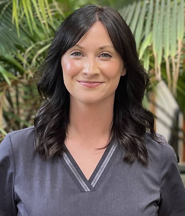 Ashley, a smiling woman with shoulder-length dark hair, is wearing grey scrubs. She is standing in front of a backdrop of lush green plants, giving the scene a fresh and welcoming atmosphere.